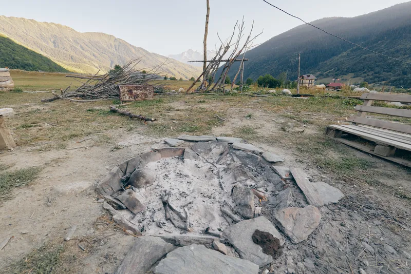 firepit ashes bench mountains rural field woodpile campfire in a quiet valley scene with charred stones, dry grass and rustic village benches near an old outdoor campsite.