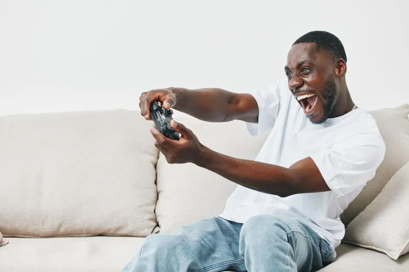 Excited young Black man playing video games on a couch, showcasing emotions of joy and engagement while gaming in a modern living room