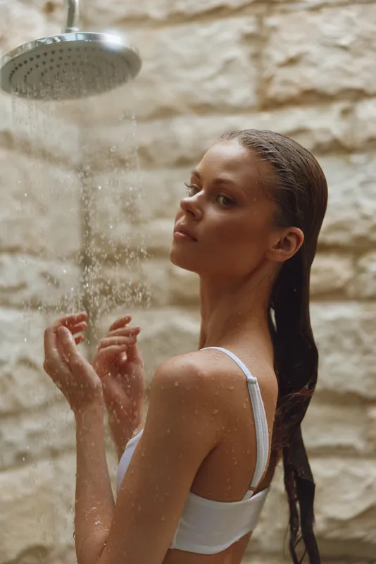Young woman taking an outdoor shower with wet hair against a natural stone wall, enjoying a refreshing and relaxing moment in warm daylight.