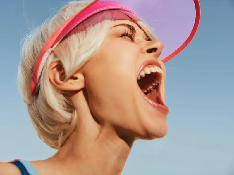 Young woman with short blonde hair wearing a pink headband, mouth open in a vivid shout, captured outdoors against a clear blue sky and bright sunlight.