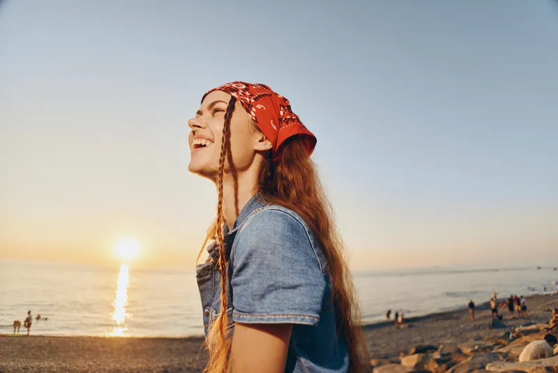 Lifestyle woman in modern boho-western denim street style smiling outdoors during sunset with warm film color tones and relaxed beach background.
