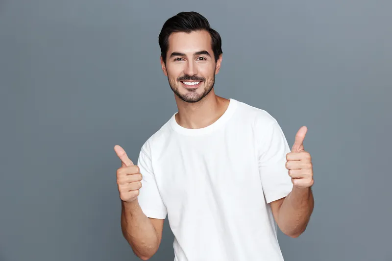 Happy smiling man showing thumbs up gesture wearing casual white t-shirt, standing against plain solid grey background. Positive attitude and cheerful expression concept.