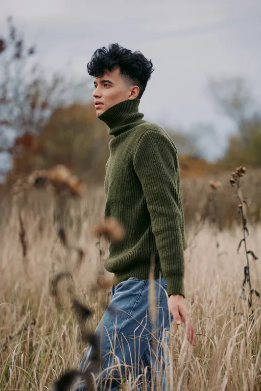 A candid, authentic portrait of a young man in an olive turtleneck sweater walking through a tall grassy meadow, captured with natural light, genuine expression and earthy tones for realism.