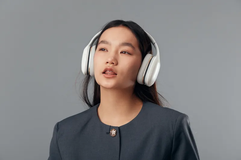 Young woman with long black hair wearing white headphones and formal blazer posing on grey background for music listening and relaxation with thoughtful expression.