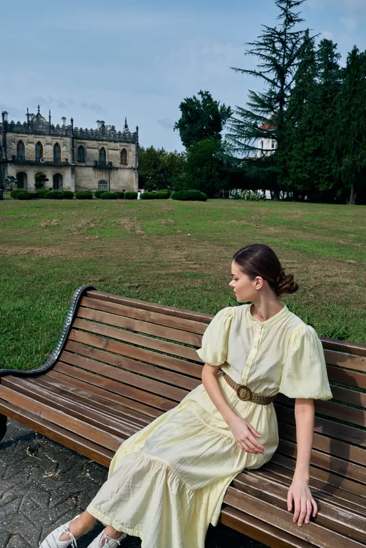 Woman in a pale yellow dress sits on a wooden bench in a park with a historic building in the background, creating a calm sunlit outdoor portrait
