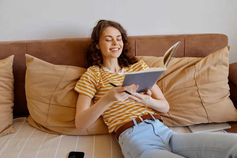 Woman relaxing at home, reading a notebook on a sofa, striped top, soft natural light, cozy living space, relaxed mood, everyday creativity, calm atmosphere
