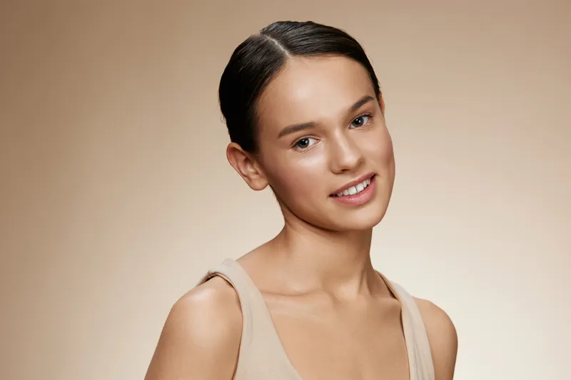 A young female model with a warm smile and natural makeup on a soft beige background, showcasing confidence and beauty in a minimalist style, perfect for health and beauty concepts