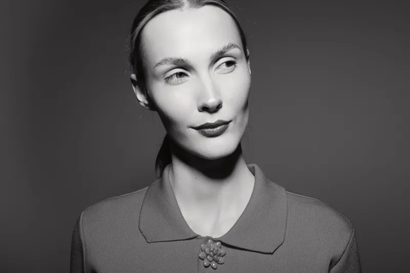 Portrait of a pale white woman with light skin and tied back hair wearing a textured top with a collar and flower detail. Studio lighting creates dramatic shadows and highlights.