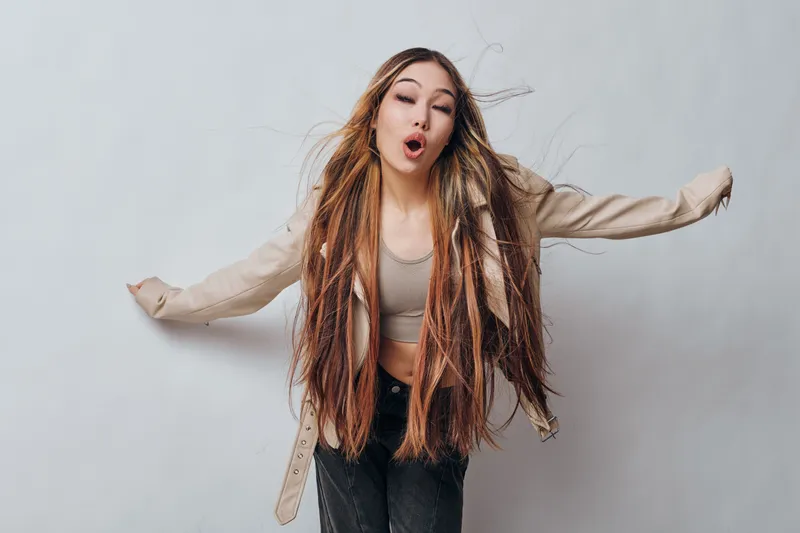 Woman with long braids and casual outfit making funny face with eyes closed and arms outstretched against plain light wall. Female fashion portrait with natural expression and hairstyle.