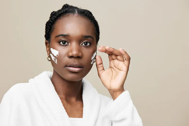 Young Black woman applying facial cream with a serene expression, wearing a white bathrobe against a neutral background, showcasing skincare and self care routine