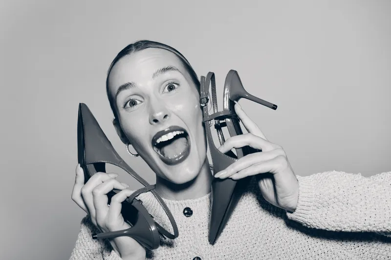 Excited Caucasian woman with light skin holding red high heel shoes near face, smiling widely on grey background. Concept for fashion, footwear, and happiness.