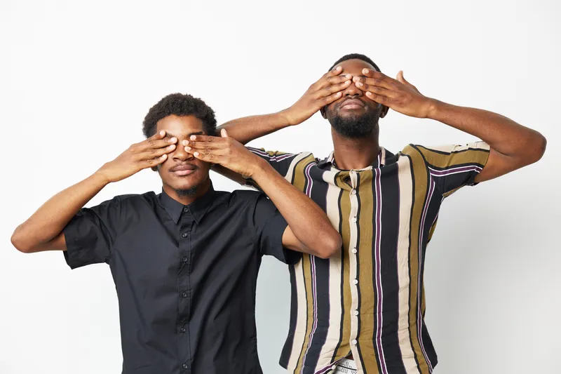 Two black men having fun, playfully covering their eyes with their hands. They express joy and camaraderie, showcasing sibling-like bonds in a bright setting.