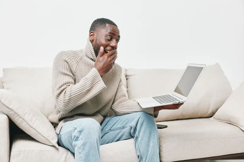 Happy young Black man with surprised expression, interacting with laptop on cozy couch, modern living room, casual style, joyful emotion, home lifestyle