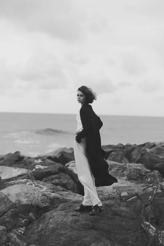 Woman standing on rugged coastal rocks, wind catching her coat as waves crash nearby, black and white scene capturing solitude, sea breeze, and distant horizon.