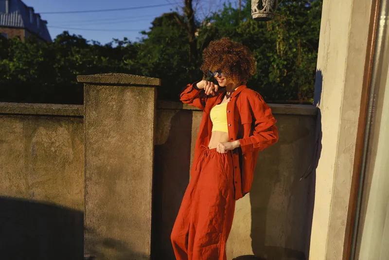 Stylish woman in red orange outfit posing on a sunny outdoor street, showcasing lifestyle and confident pose, urban backdrop with warm light, fashionable yet relaxed mood and natural expression