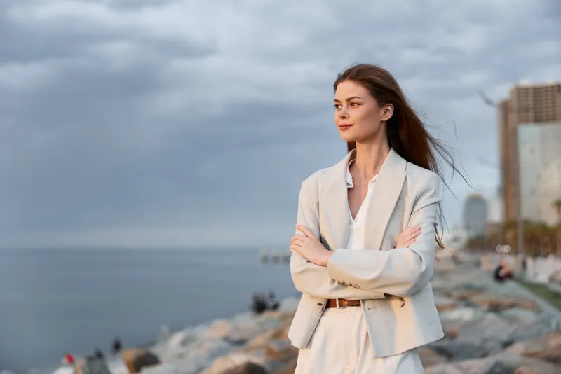 confident business woman in white suit standing by the rocky sea shore with arms crossed and thoughtful expression under cloudy sky