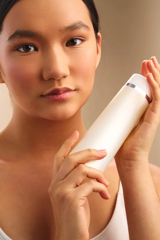 Asian woman holds a smooth bottle near her face in a soft, warm setting. Her calm expression and subtle smile convey beauty, care, and confident wellness during a skincare moment.