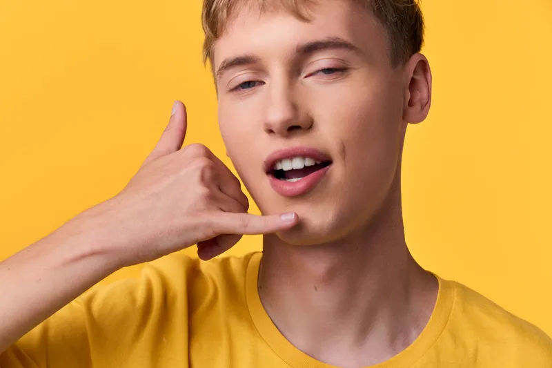 Hand gesture and playful phone pose for casual communication and modern style, featuring a smiling young man in a yellow shirt against a bright yellow background