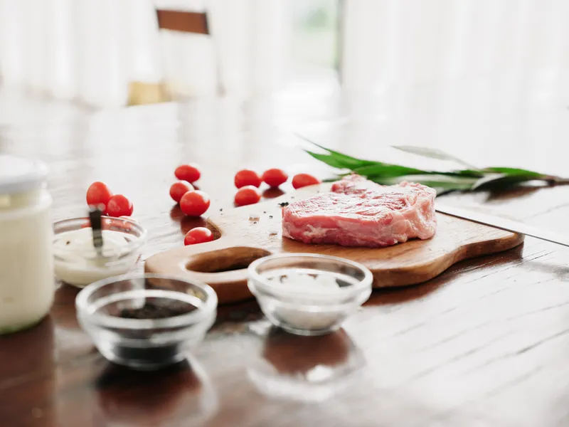 Стильная кухня, на деревянном столе лежит кусок свежего мяса со специями, помидоры и зелень, свет от окна