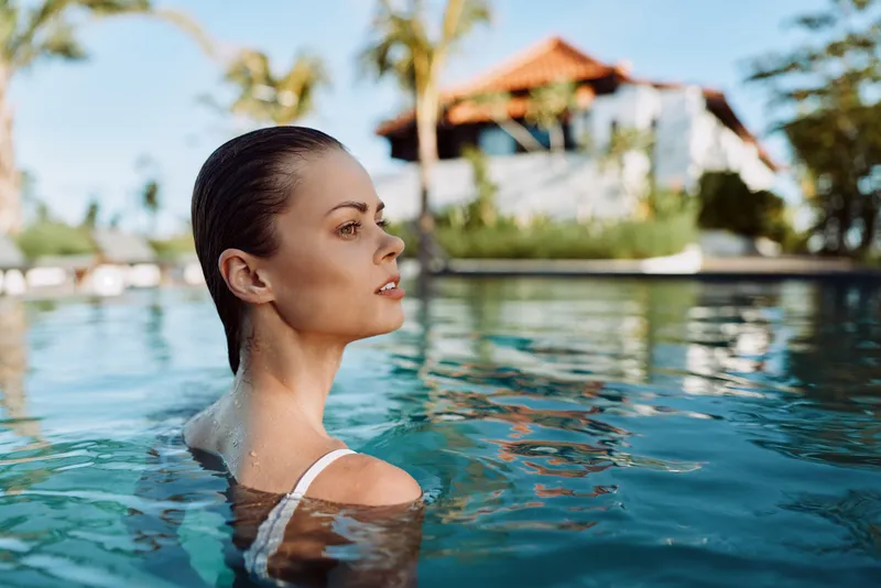 Relaxed woman in a sunlit pool, head tilted back, calm smile, tropical resort backdrop, clear water, soft breeze, casual swimwear, candid outdoor moment, natural colors, vacation mood