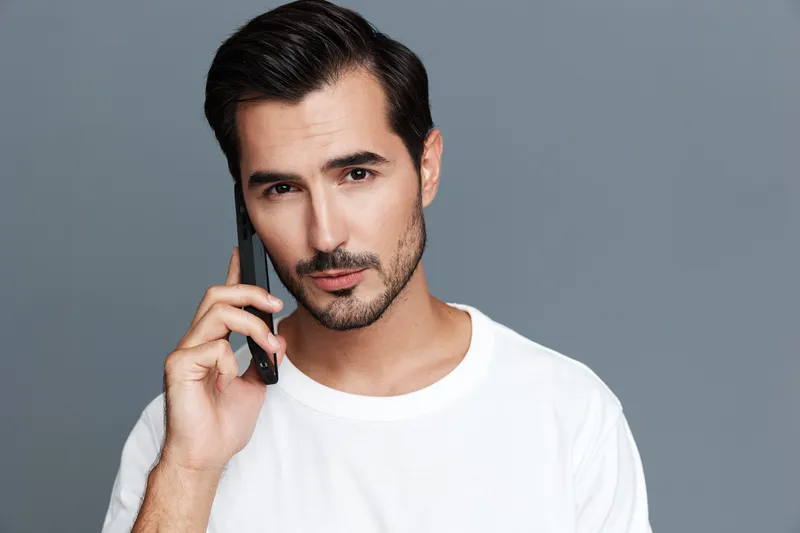 Handsome man talking on mobile phone, dressed in casual white t-shirt, with serious expression, isolated on solid gray background. Lifestyle concept.