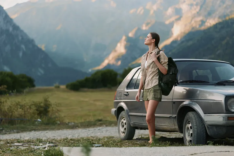 Young woman with backpack enjoying a mountain landscape beside a parked car, gazing into the distance. A carefree outdoor lifestyle captures adventure, travel, and nature in warm, soft light on