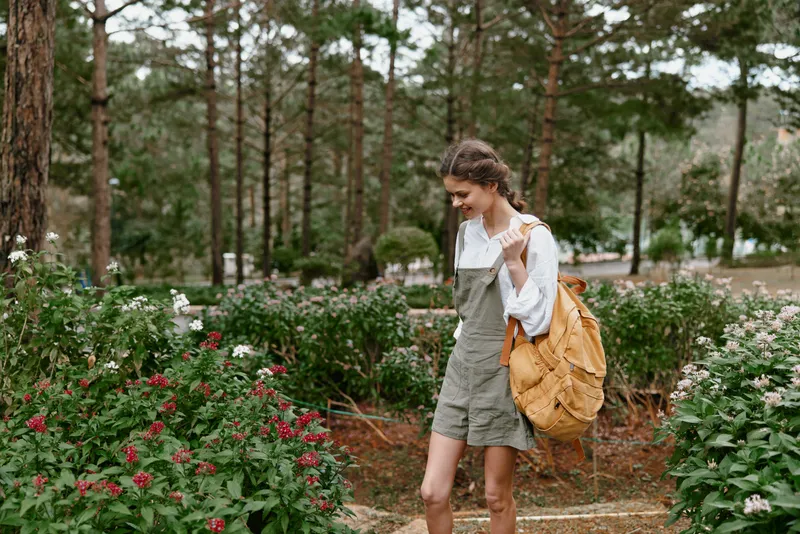 Young woman wearing comfortable overalls and carrying a backpack while walking through a vibrant garden, illustrating a joyful outdoor experience in nature