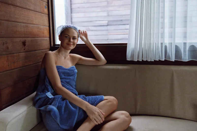 Young woman wrapped in a blue towel sitting comfortably on a beige sofa by a bright window, relaxed and peaceful in a cozy wooden interior.