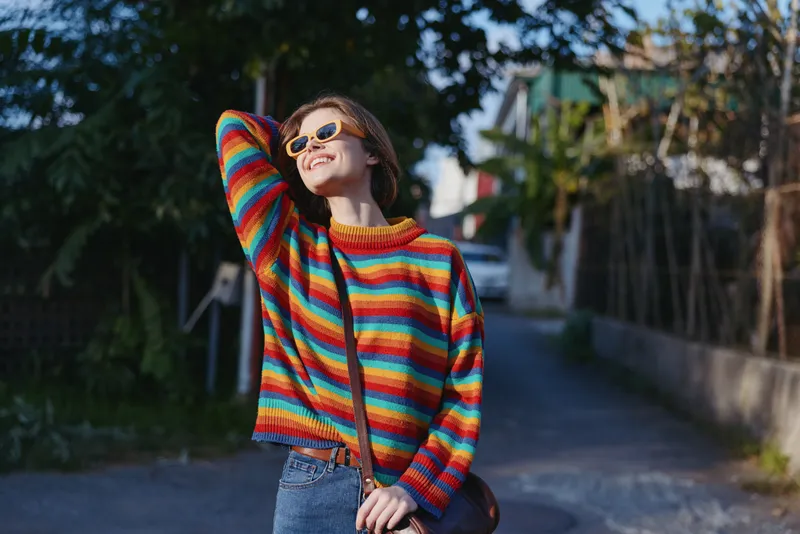 young woman striped sweater smiling sunglasses casual outfit urban street, 20s model leaning back in rainbow knit sweater enjoying sunny travel lifestyle and relaxed joyful mood