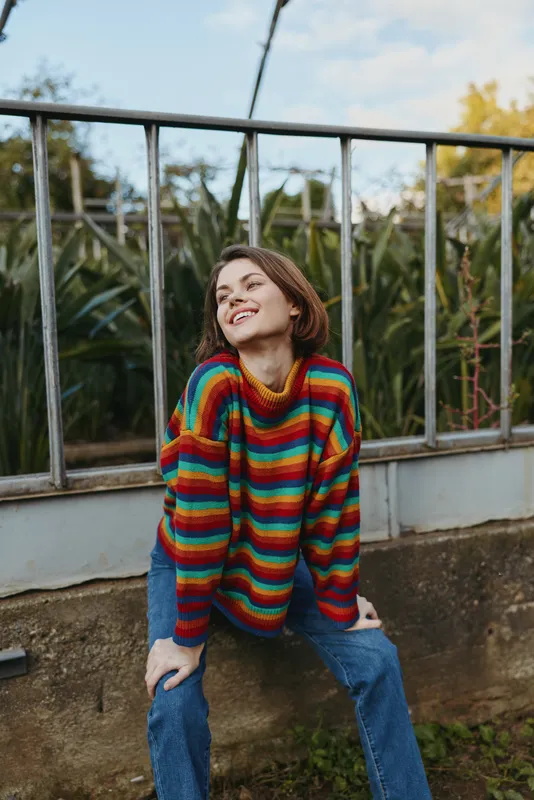Woman in a rainbow striped sweater leaning on a concrete wall, smiling young adult with short hair in a casual outfit and jeans, outdoor urban garden scene with relaxed pose.