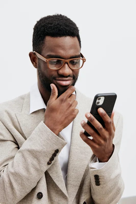 Thoughtful man wearing glasses in beige blazer holding smartphone looking at screen isolated on plain white background studio portrait. People lifestyle concept