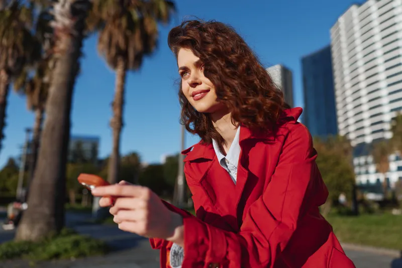 Young woman with curly hair wearing red jacket points finger outdoors in urban park with palm trees and modern buildings in background on sunny day.