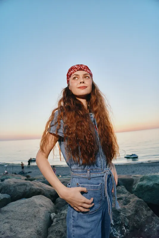 Lifestyle woman in modern boho-western denim street style stands confidently by the rocky shore at sunset, showcasing film color tones and casual outdoor fashion.