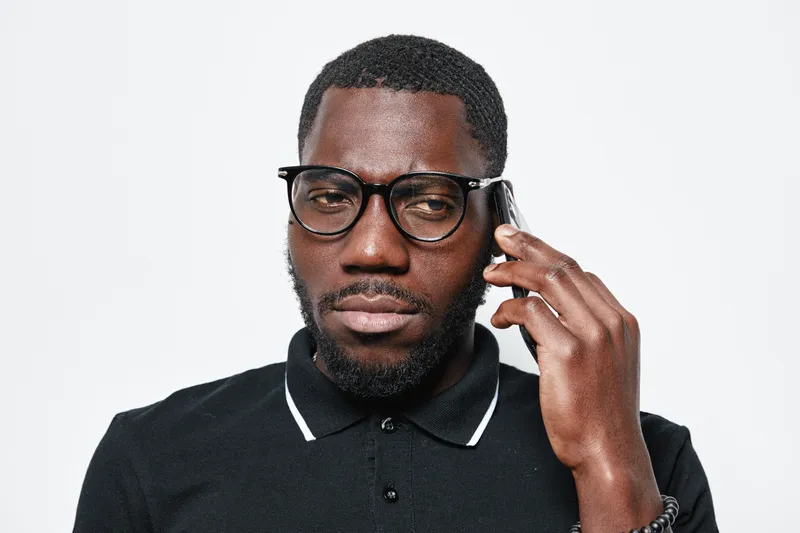 Black man with dark skin and beard wearing glasses and black polo shirt talking on smartphone isolated on white background. Serious expression and casual style portrait.