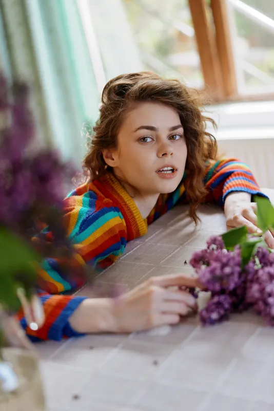 Young woman with a colorful striped sweater leans over a sunlit windowsill, reaching toward a bouquet of lilacs while a gentle, thoughtful smile lights her face.
