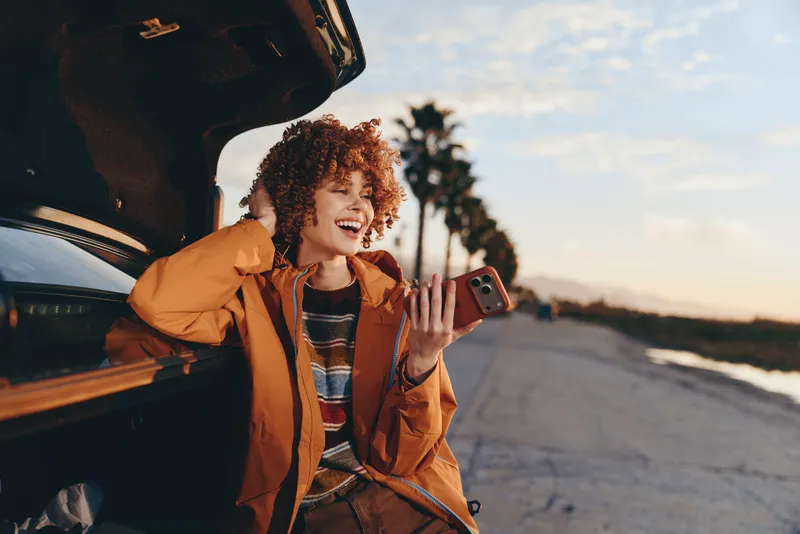 Woman lifestyle, curly haired woman laughing in orange jacket beside open car door, holding phone in hand, road and sunset scenery, warm evening light, travel outdoors