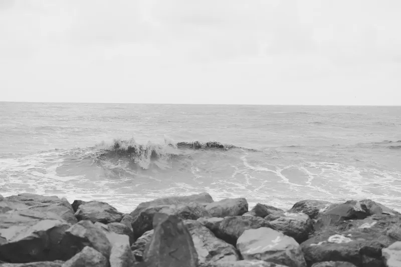 Monochrome seascape with rocky shoreline and rolling waves, calm sea surface, rugged coast, distant horizon, overcast sky, and textured stones in the foreground, conveying solitude and natural mood