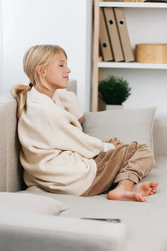 A young girl sits barefoot on a soft sofa, wearing a cream knit sweater and cozy trousers, posing in a peaceful, intimate moment of quiet comfort.