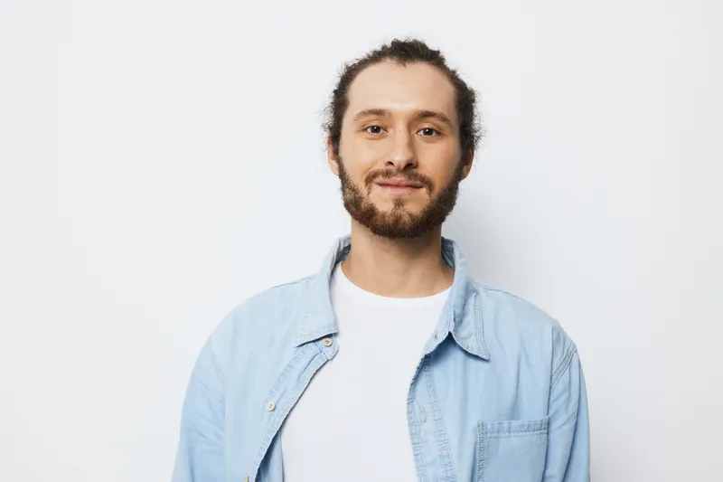 Young man smiling confidently in a light blue shirt against a plain white background, showcasing a positive and approachable demeanor, ideal for lifestyle and portrait photography