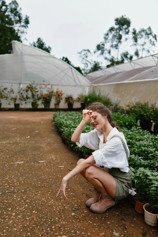Smiling young woman in green shorts and white shirt in a greenhouse, showcasing a joyful connection with nature and gardening, surrounded by lush plants and flowers