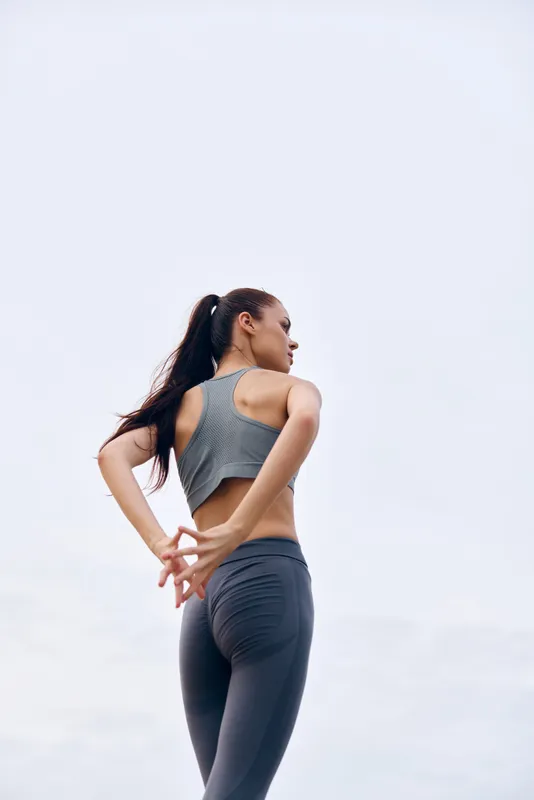 Athletic young woman stretching outdoors, dressed in sportswear, showcasing determination and focus against a serene grey sky Ideal for fitness and wellness themes