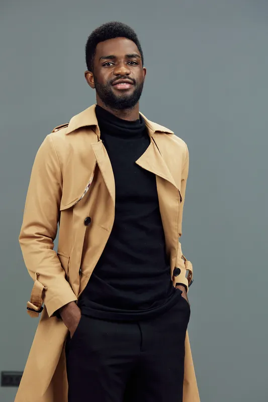 Stylish young Black man in a tan trench coat and black turtleneck, exuding confidence against a minimalist gray background, ideal for fashion concepts