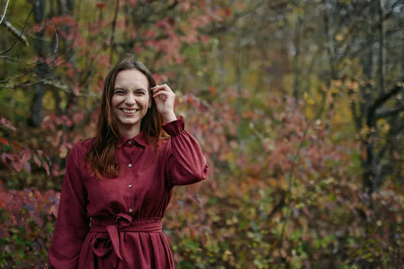 A candid portrait of a smiling woman in a burgundy dress stands in autumn woods, conveying authenticity and credibility through natural light and unposed expression.