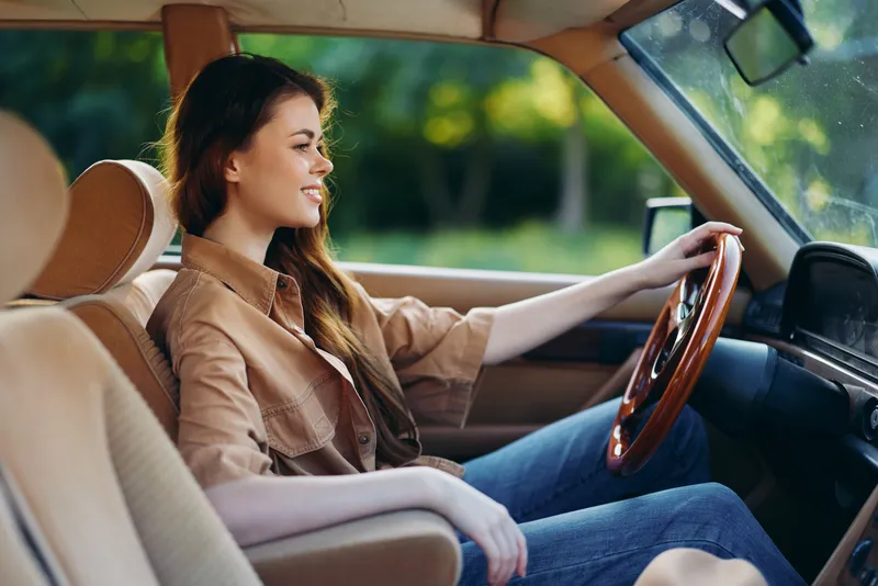 Stylish young woman driving a vintage car, wearing a brown shirt and jeans, with a confident smile, capturing a sense of freedom and adventure.