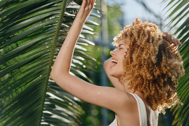 smiling woman with curly hair enjoying tropical sunlight near palm leaves, natural beauty, summer mood, outdoor lifestyle scene in warm daylight.