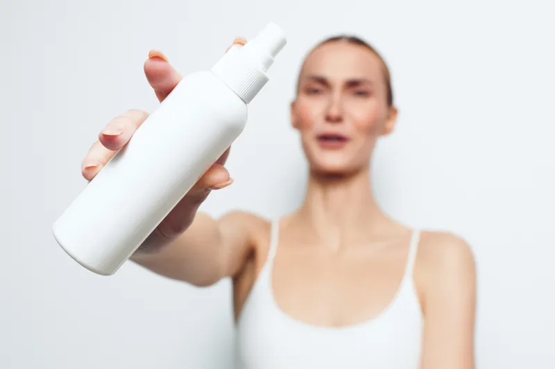 Blurred woman in white top holds a skincare spray bottle, showing beauty hygiene, cosmetic treatment and spa wellness. Soft studio background suggests calm self care for daily skin care.
