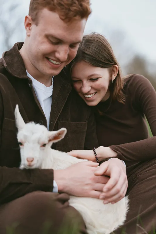Couple outdoors cuddling a goat kid, warm laughter, gentle light, relaxed posture, natural colors, rustic backdrop, casual outfits, affectionate moment, nature setting