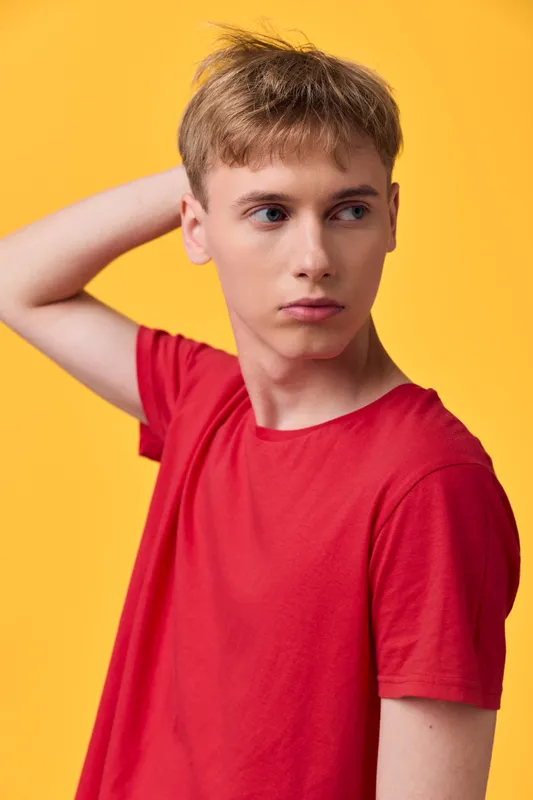 Thoughtful boy in a bright red shirt stands against a vivid yellow backdrop, casual pose with hand behind neck, studio lifestyle image for fashion or branding