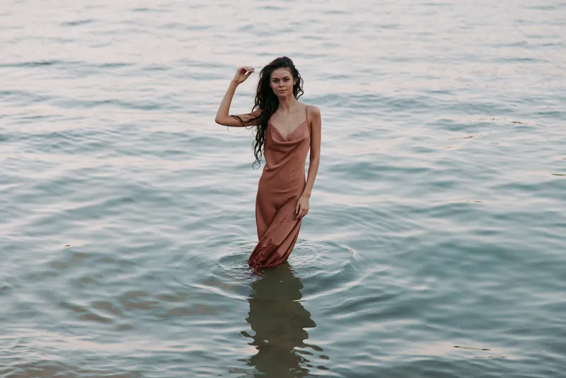 A woman stands waist deep in calm water wearing a soft brown dress, creating a serene mood and graceful pose as gentle ripples surround her.