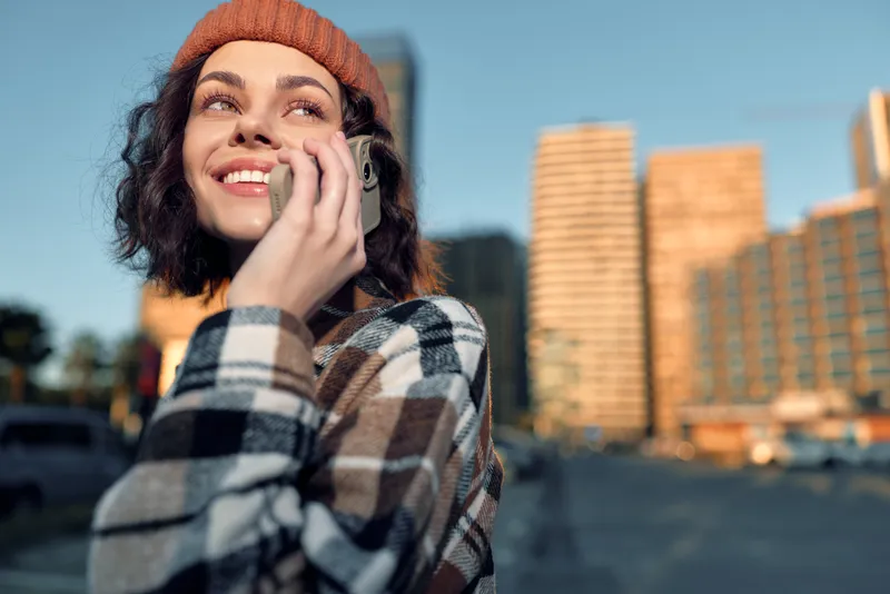 Woman phone smile city beanie coat golden hour: candid lifestyle portrait of a smiling young woman on a mobile call in urban street at sunset, authenticity and emotional storytelling vibe.